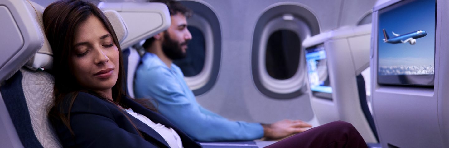 Nahaufnahme einer Frau, die leicht zurückgelehnt im Flugzeug schläft, während der Mann neben ihr das Bordprogramm auf dem Bildschirm im Vordersitz anschaut.