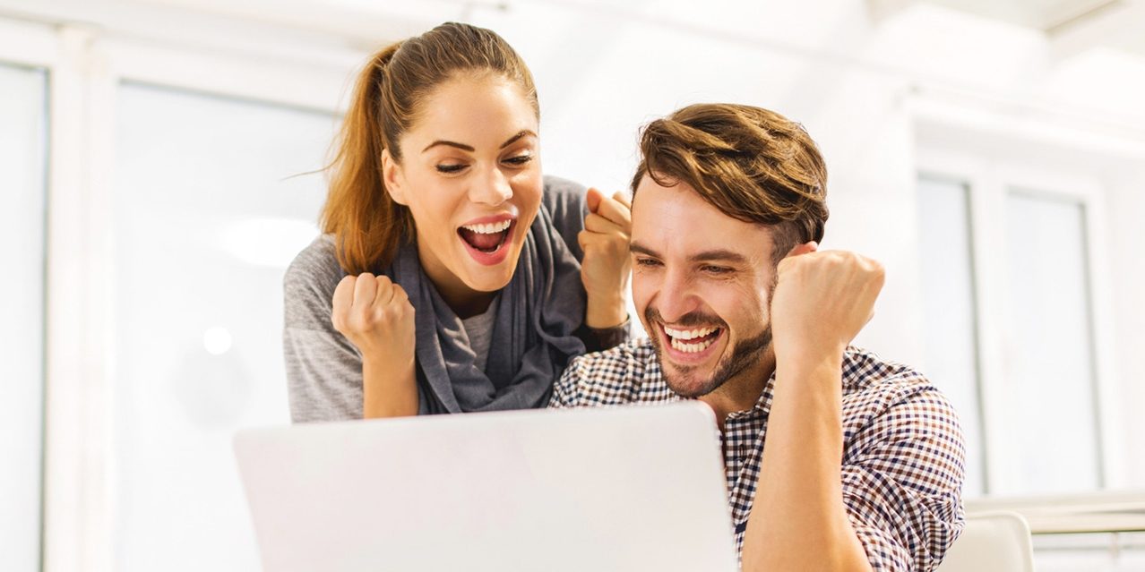 Two people sitting at a table with a laptop, both raising fists in a celebratory gesture.