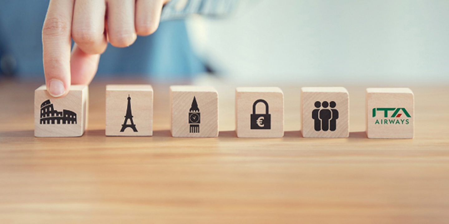 A hand picking up a block from a row of wooden blocks with travel icons and the ITA Airways logo.