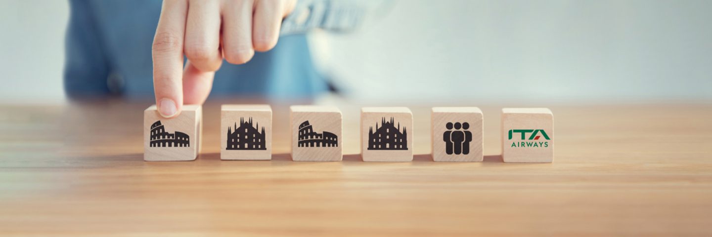 A hand picking up a block from a row of wooden blocks with travel icons and the ITA Airways logo.