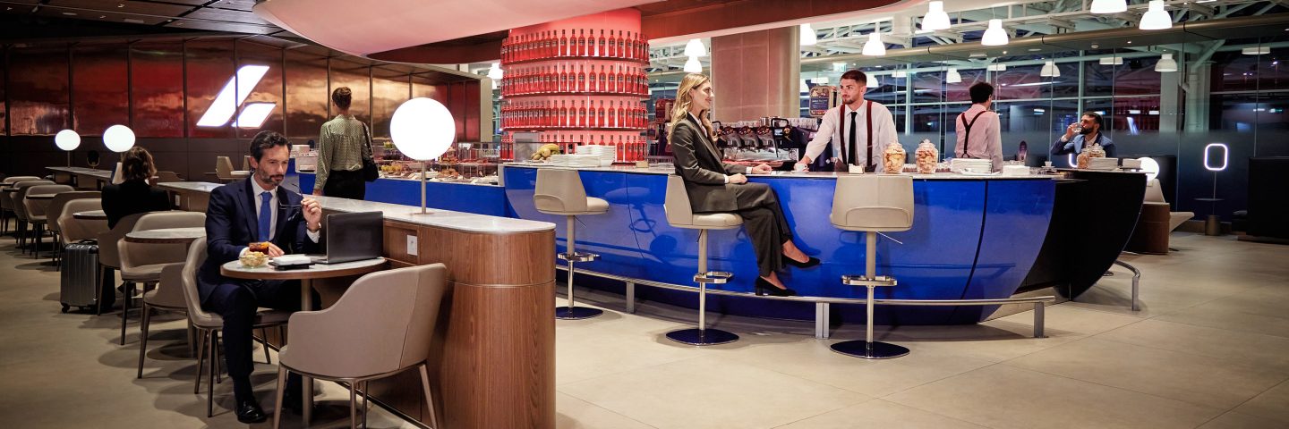 An airport lounge with a bar area and passengers seated at tables.