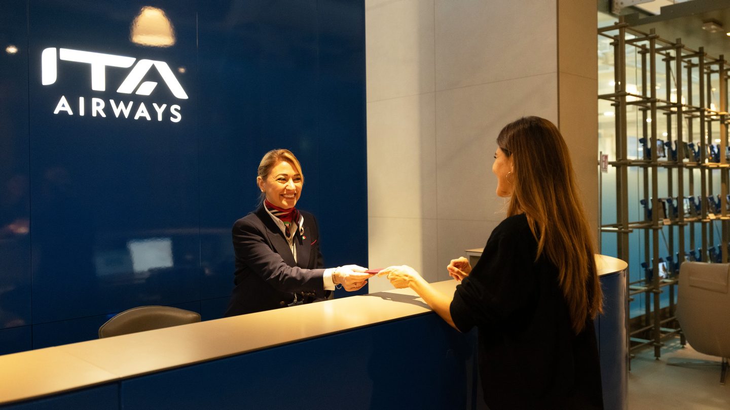 Lounge reception desk with ITA Airways staff and a passenger.