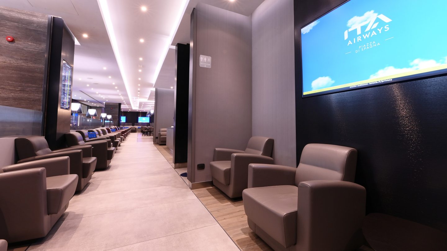 Couloir d’un salon avec des fauteuils et un écran affichant « ITA Airways » fixé au mur.