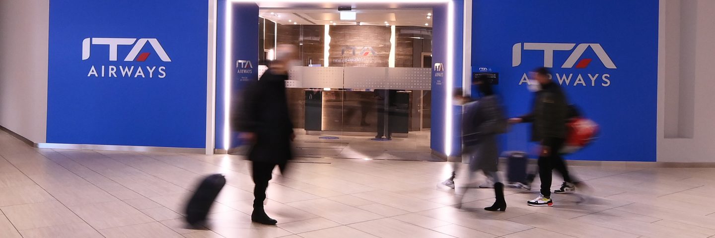 Entrée du salon avec des murs bleus ITA Airways et des personnes passant devant.