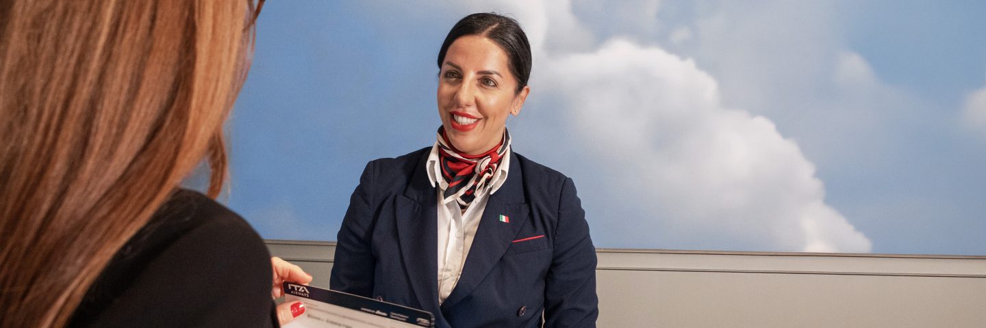 Ein Mitglied des ITA Airways Personals sitzt lächelnd am Schalter und nimmt ein Dokument von einem Passagier entgegen.