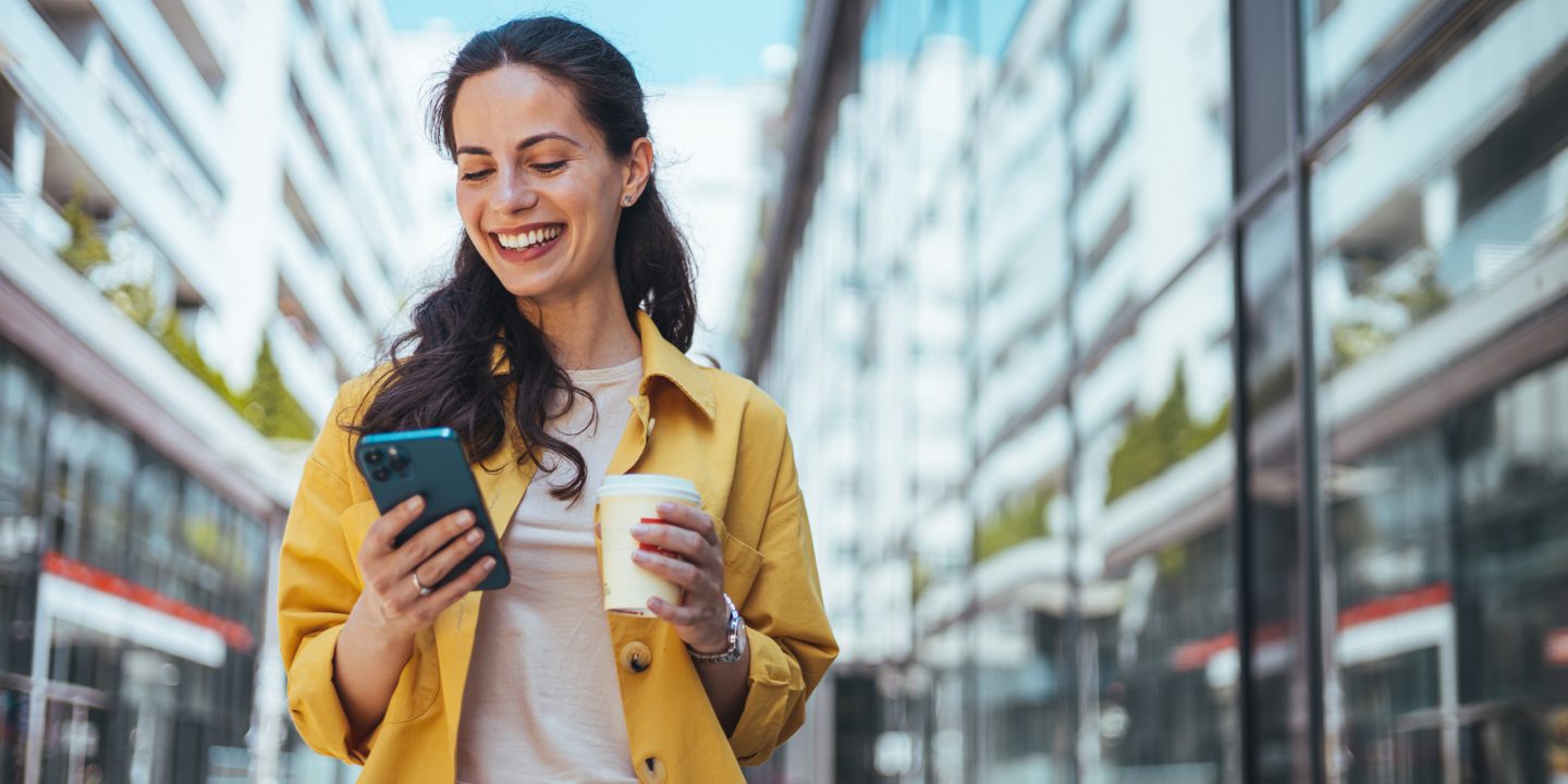 Una mujer con un abrigo amarillo sonríe mientras usa su teléfono con una mano y sostiene una taza de café con la otra.