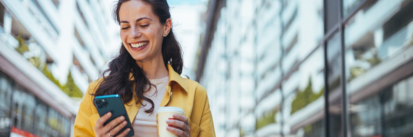 Une femme portant un manteau jaune sourit, son téléphone dans une main et une tasse de café dans l’autre.