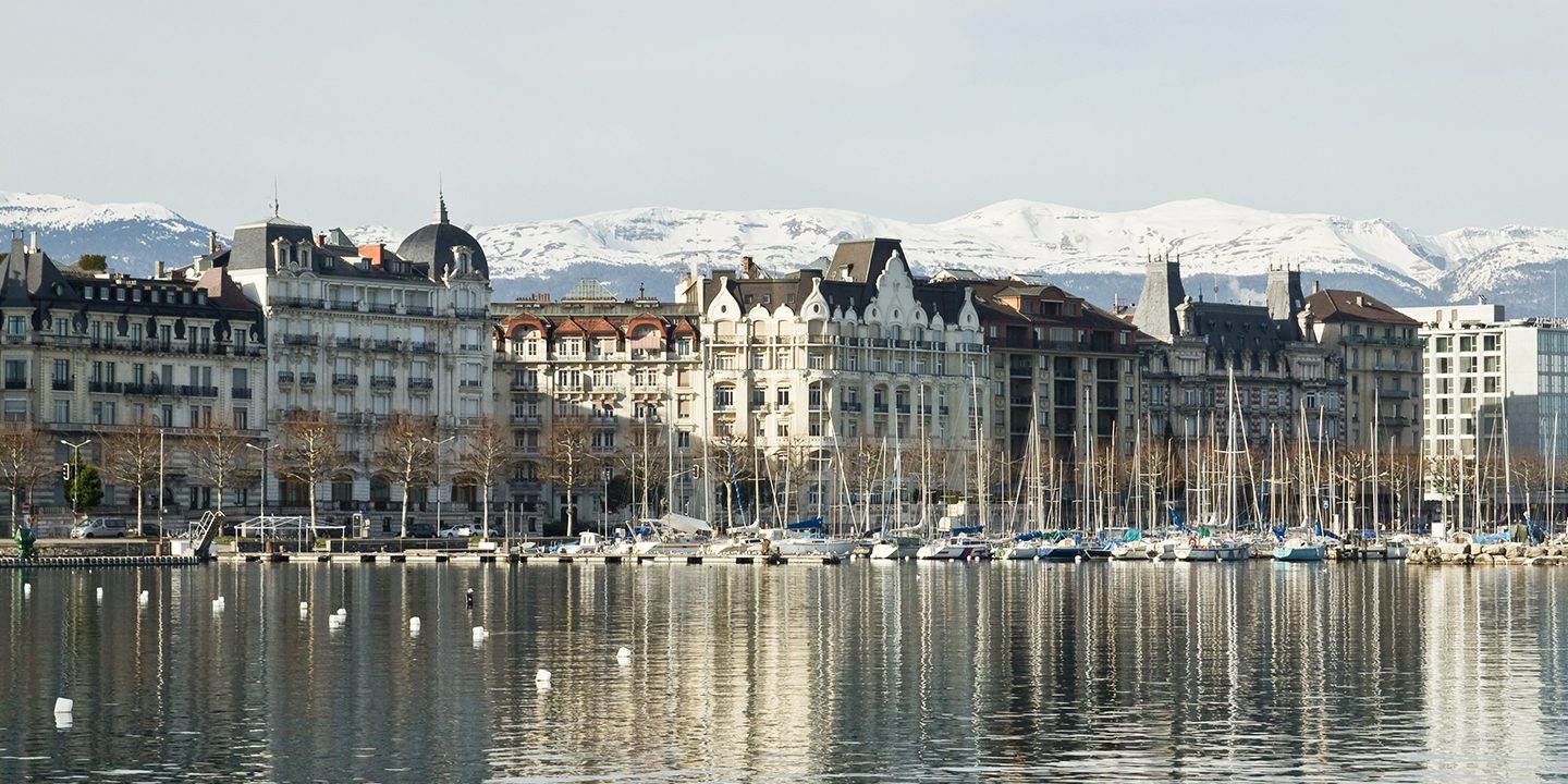 Buildings along the Geneva lakefront, reflected in the water, with snow-capped mountains in the background.