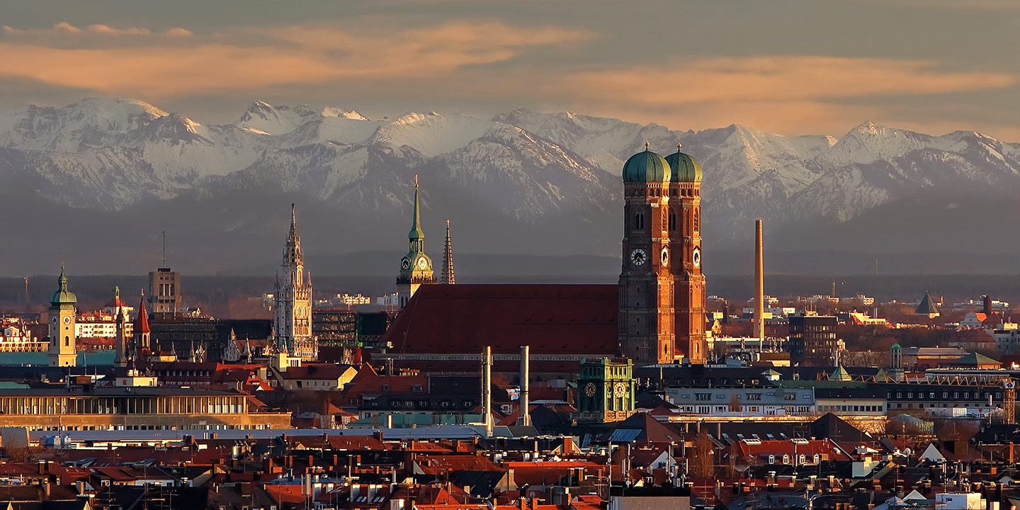 The Munich skyline with the Frauenkirche and the snow-capped Alps at sunset.