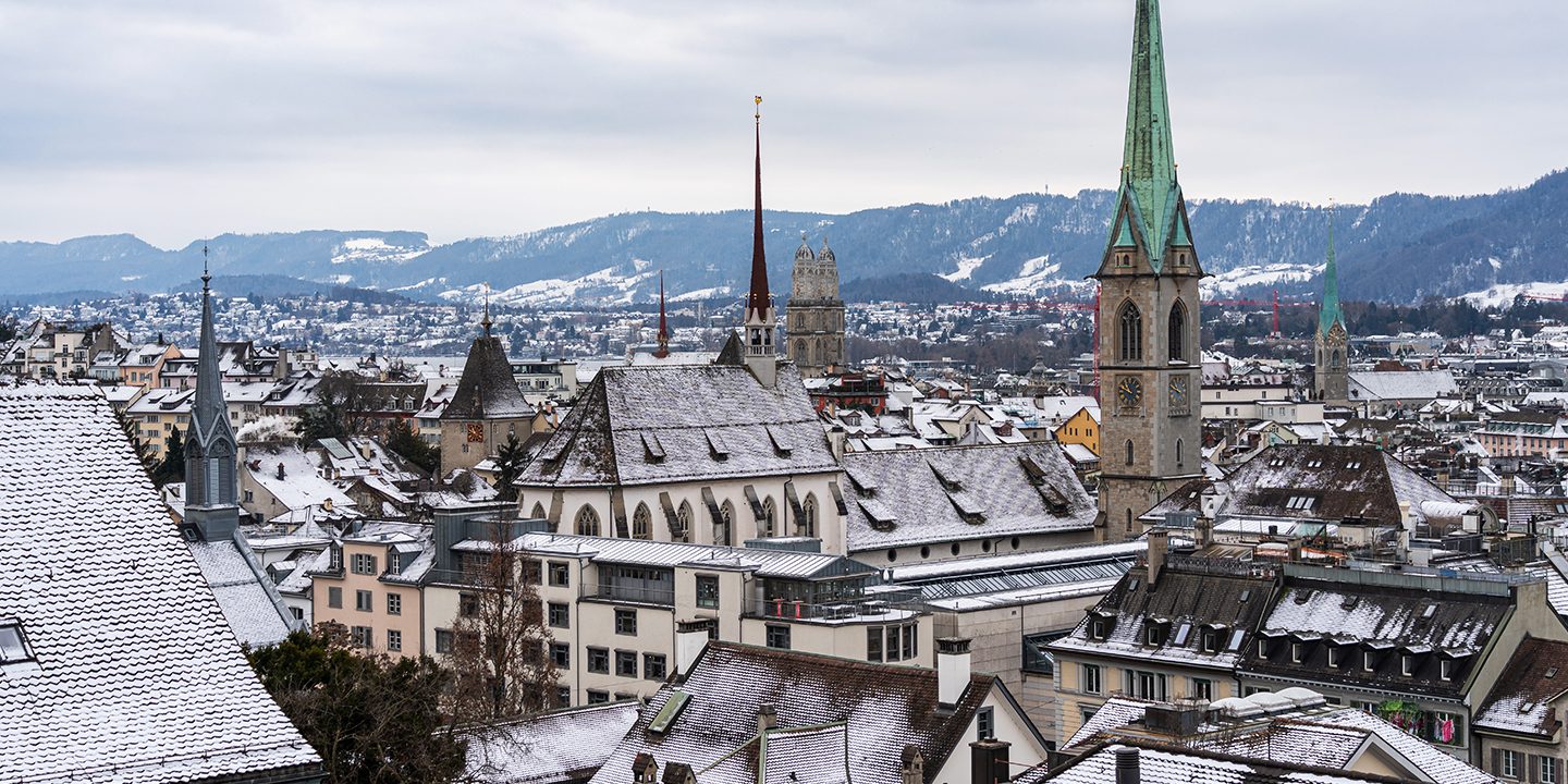 Snowy, urban Zurich landscape with bell towers and hills in the background.