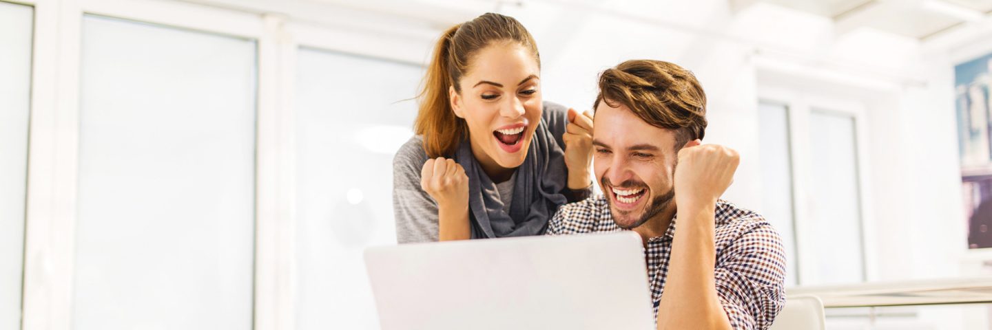 Two people sitting at a table with a laptop, both raising their fists in a gesture of victory.