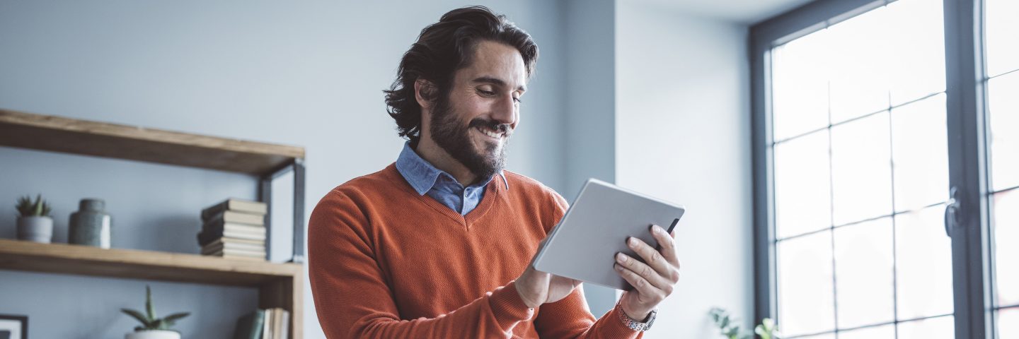 Un homme sourit en utilisant une tablette et en s’appuyant sur un bureau.
