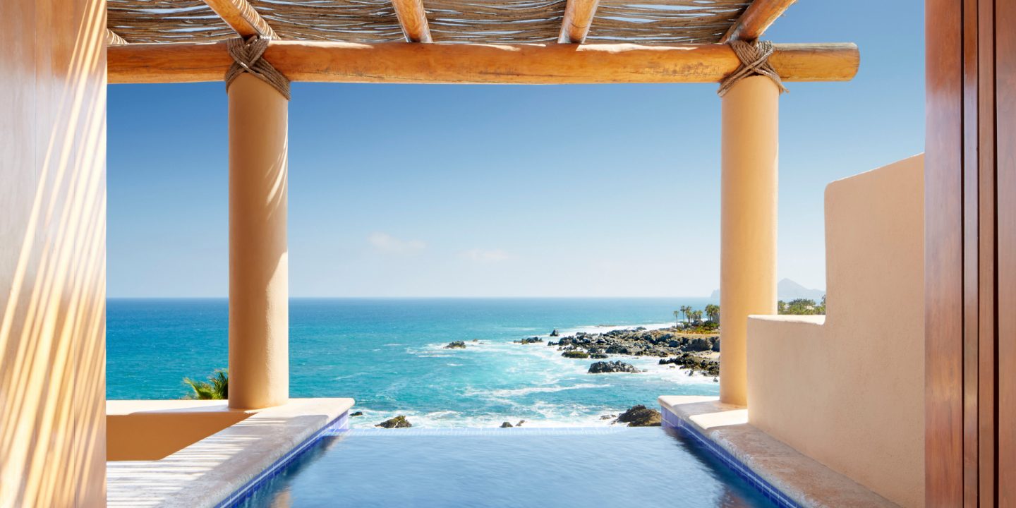 View from a terrace with a pool overlooking a rocky coastline and the blue ocean under a clear sky.