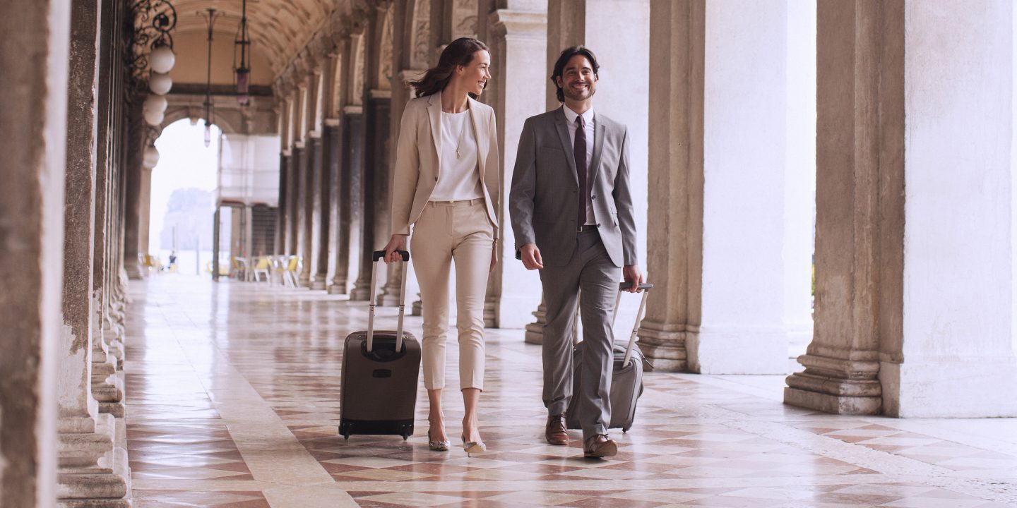Two people in business attire walking under arcades in Rome with suitcases.