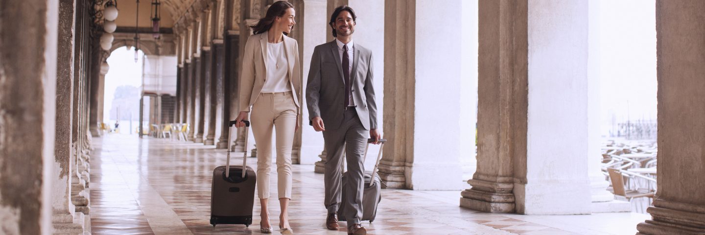 Two people in business attire walking under arcades in Rome with suitcases.