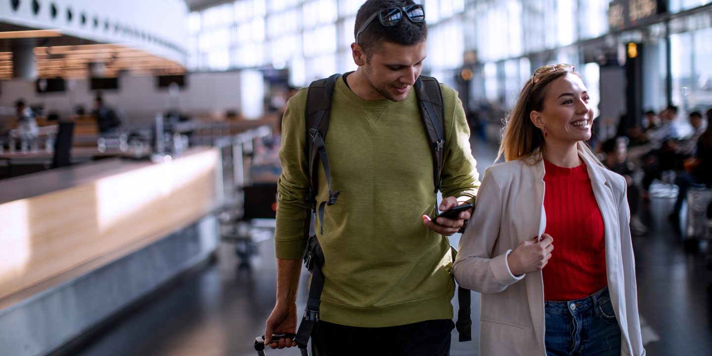 Two passengers walking through the airport, one checking their smartphone.