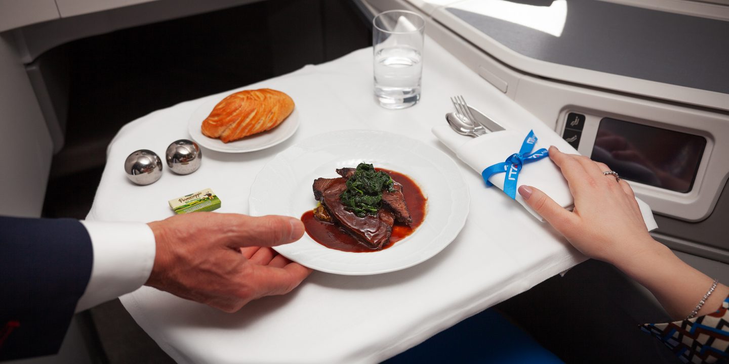 A person serves a beef dish with vegetables, a bread roll, butter, salt, pepper, and water.