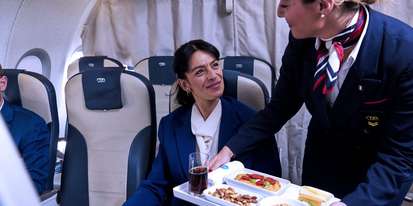 A woman in a blue uniform serves food to a woman seated on an airplane.