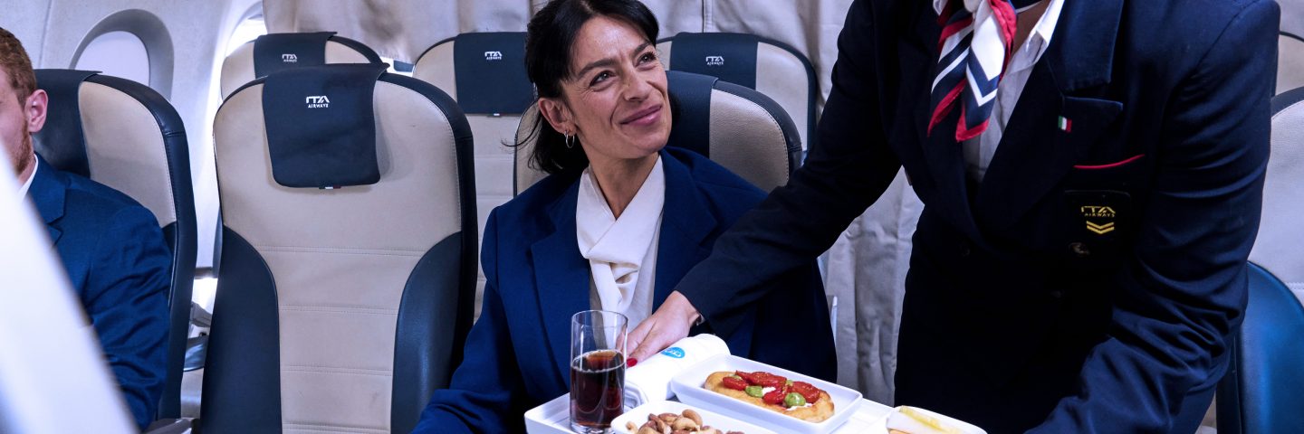 A woman in a blue uniform serves food to a woman seated on an airplane