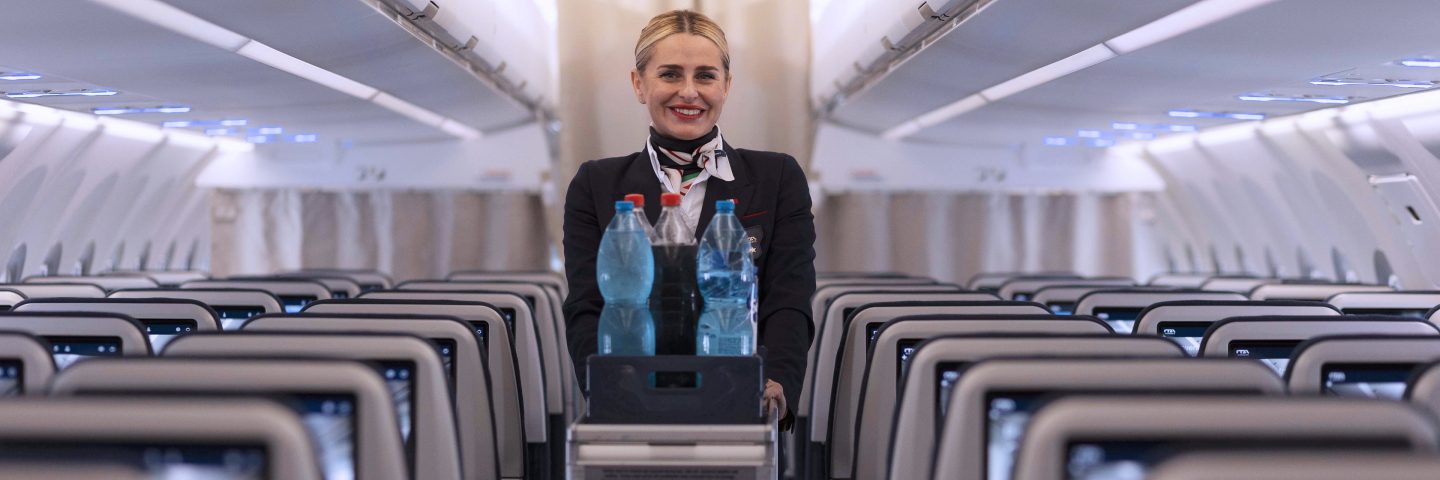 Assistente di volo nel corridoio di un aereo ITA Airways con carrello d’acqua.