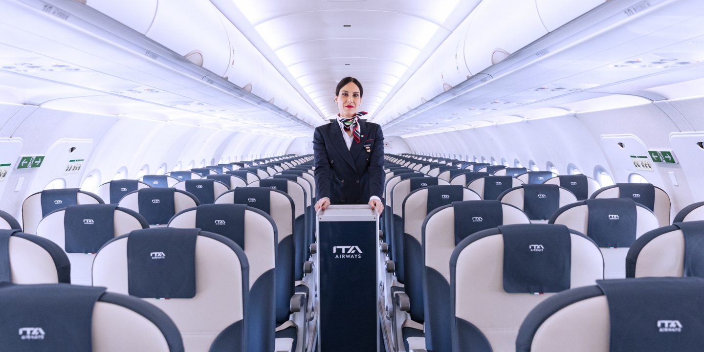 A woman standing in the aisle of an aircraft, with seats visible on the sides.
