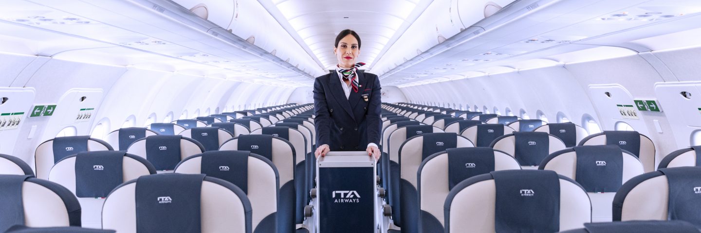 Una donna in piedi nel corridoio di un aereo, con sedili visibili ai lati.
