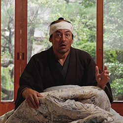 Homem vestido de samurai sentado no chão em uma sala japonesa tradicional com uma expressão de surpresa.