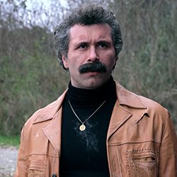 Um homem com um bigode grosso, usando uma jaqueta de couro e uma gola alta preta.