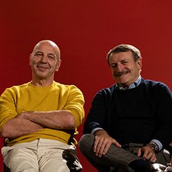 Dois homens sorridentes sentados juntos contra um fundo vermelho sólido.