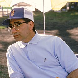 Eugene Levy usando uma camisa polo, óculos e um boné de malha sob um guarda-chuva.