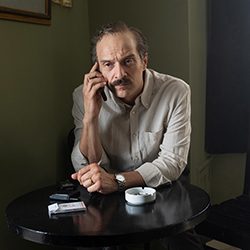 Um homem de bigode falando ao telefone em uma mesa com um cinzeiro.