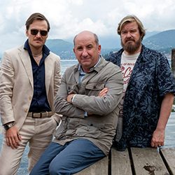 Três homens juntos em um píer de madeira com água ao fundo.