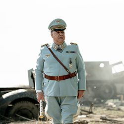 Homem de uniforme militar histórico azul-claro, com medalhas, em um campo desolado e cheio de detritos.