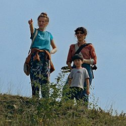 Duas mulheres e um menino em pé em uma colina gramada sob um céu azul, com uma mulher apontando para a distância.