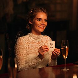 Samara Weaving em um vestido de noiva de renda sorrindo enquanto segura um cartão com “esconde-esconde”.