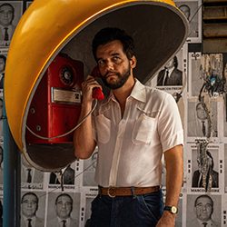 Homem barbudo com uma camisa branca falando ao telefone em uma cabine telefônica amarela vintage.
