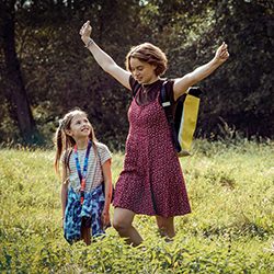 Mulher caminhando com os braços abertos ao lado de uma menina em um campo gramado.