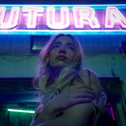 Mulher jovem com cabelo loiro posando sob uma placa de neon rosa.