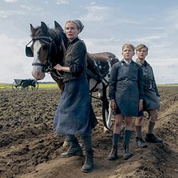 Kirsten Dunst com um cavalo e dois meninos em pé em um campo enlameado.