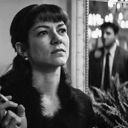 Olivia Ross segurando um cigarro com Janula Burger refletido em um espelho.