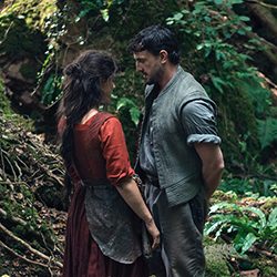 Um homem e uma mulher com roupas de época, frente a frente em uma floresta.