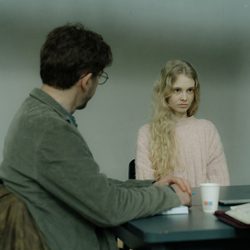 Homem conversando com uma mulher loira de suéter rosa do outro lado de uma mesa.