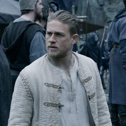 Um homem com o cabelo penteado para trás usa uma jaqueta acolchoada bege ao ar livre.