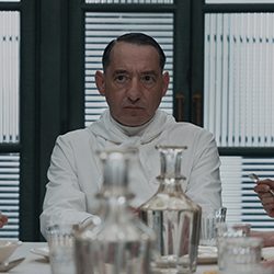 Um homem austero em uniforme branco sentado à mesa de jantar atrás de decantadores de água.