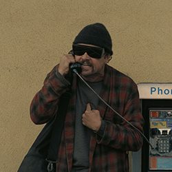 Um homem usando um gorro preto, óculos escuros e uma jaqueta xadrez vermelha e preta falando em um orelhão.