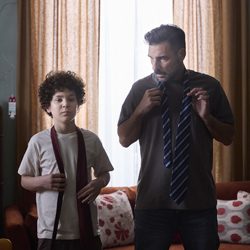 Um homem e um menino ajustam as gravatas no pescoço, dentro de um quarto.