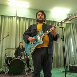 Homem tocando uma guitarra elétrica azul-petróleo no palco, com um baterista atrás dele.