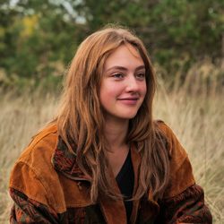Jovem sorridente com cabelo marrom claro sentada ao ar livre em um campo.
