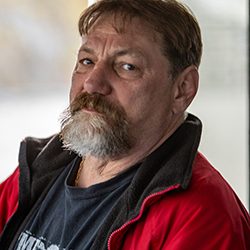 Homem de meia-idade com bigode e barba veste uma jaqueta vermelha.