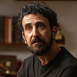 Close-up de um homem com cabelo escuro e cacheado e barba olhando pensativamente para o lado.